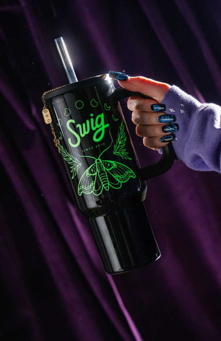 A hand in a purple sleeve holds the glowing moth side of the Witching Hour tumbler against purple drapes.