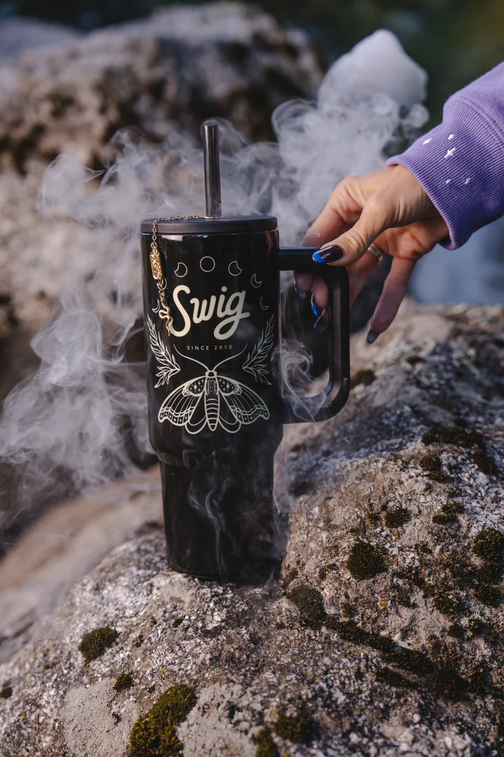 The black moth-design Witching Hour tumbler rests on rough stone surrounded by mist, with a hand on its handle.