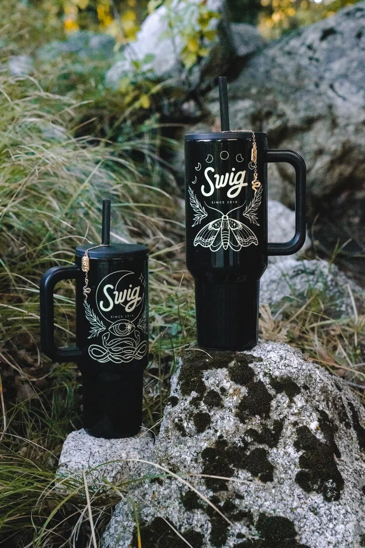 Two black Witching Hour tumblers on woodland rocks, showing the moon and serpent artwork on the left and moth artwork on the right.